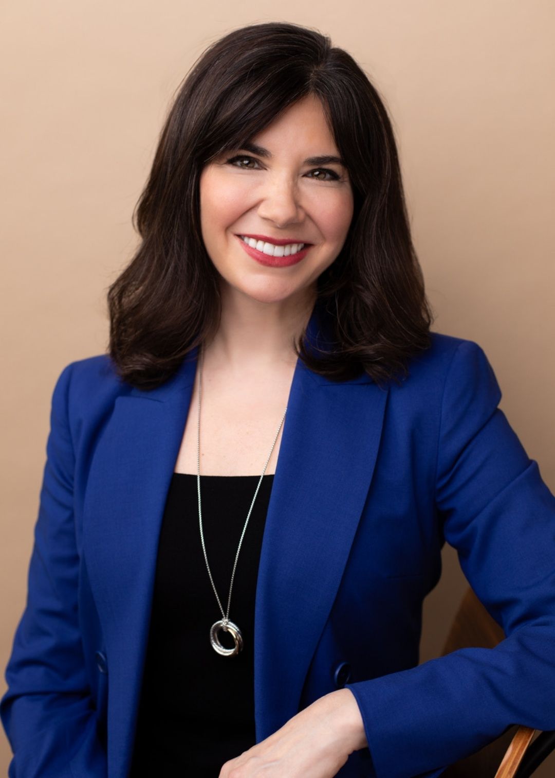 Woman with shoulder-length dark hair wearing a blue blazer and a long necklace, smiling and seated against a neutral background—perfect for professional testimonials.